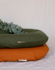 Two Bubba Clouds Are Pictured Against A White Sheet. The Orange Earth Bubba Cloud Linen Lounger Is On The Bottom And The Forest Green Bubba Cloud Linen Lounger Is On The Top. On Top Of The Green Bubba Cloud Lounger Is Eucalyptus.