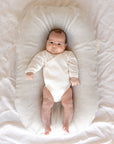 Baby Pictured In The Bubba Cloud Sand Dune Baby Lounger Wearing A White Long Sleeve White Body Suit Looking At The Camera. The Background Is A White Sheet.