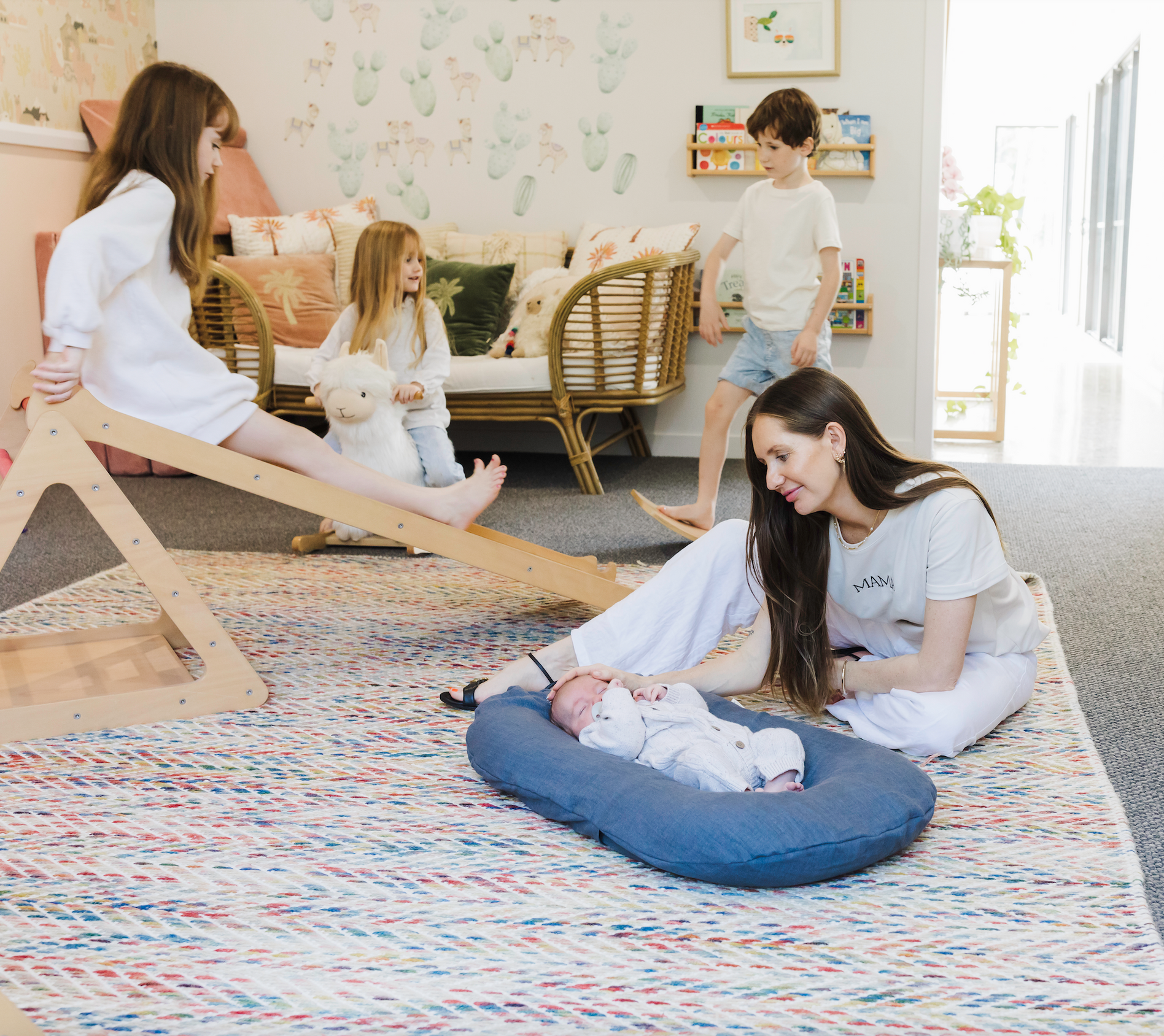 Mum With Four Of Her Children In A Kids Playroom, Looking At Her Baby Laying In A Bubba Cloud Linen Blue Lounger Whilst The Three Other Children Are Playing 