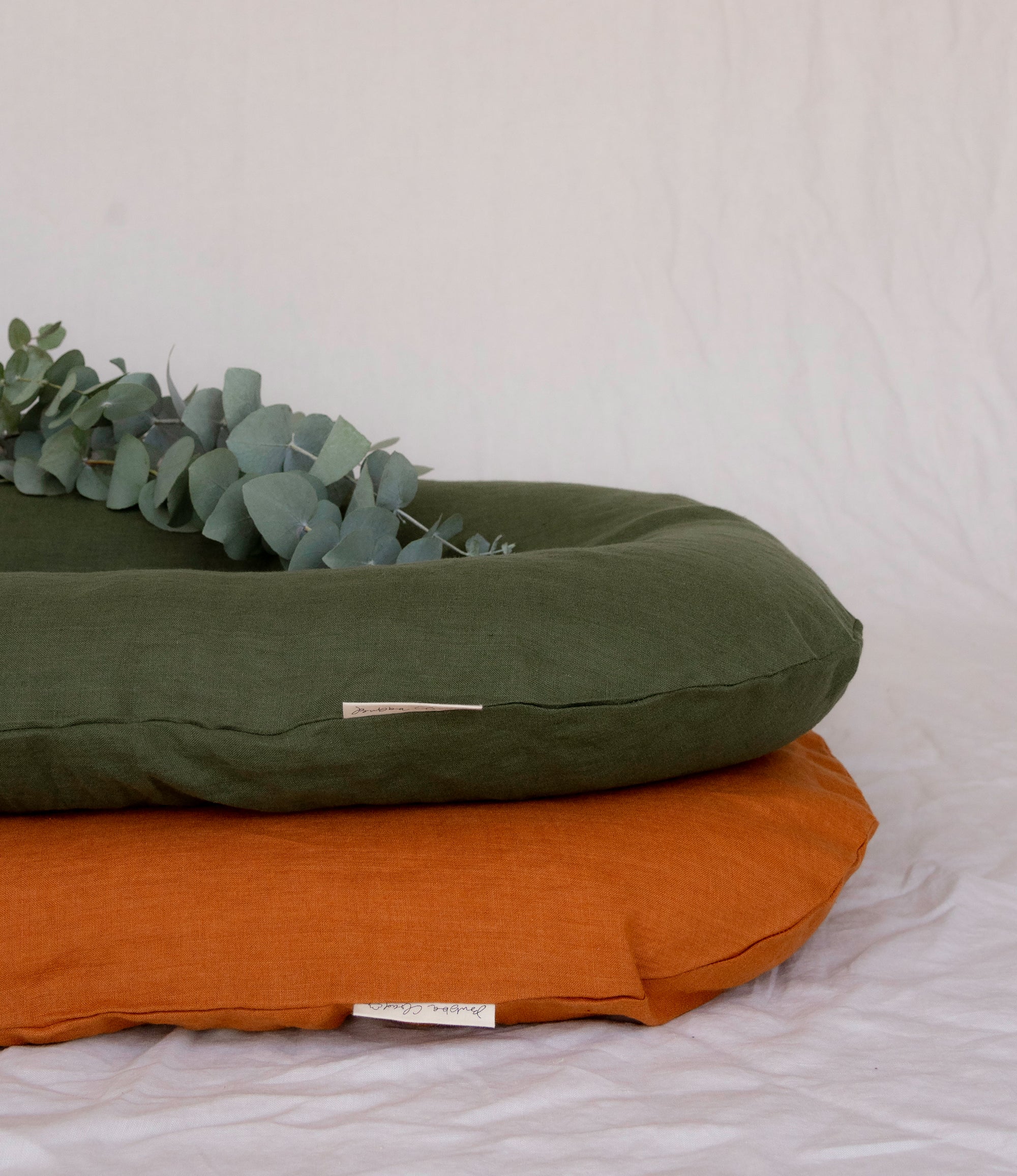 Two Bubba Clouds Are Pictured Against A White Sheet. The Orange Earth Bubba Cloud Linen Lounger Is On The Bottom And The Forest Green Bubba Cloud Linen Lounger Is On The Top. On Top Of The Green Bubba Cloud Lounger Is Eucalyptus. 