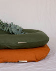 Two Bubba Clouds Are Pictured Against A White Sheet. The Orange Earth Bubba Cloud Linen Lounger Is On The Bottom And The Forest Green Bubba Cloud Linen Lounger Is On The Top. On Top Of The Green Bubba Cloud Lounger Is Eucalyptus. 