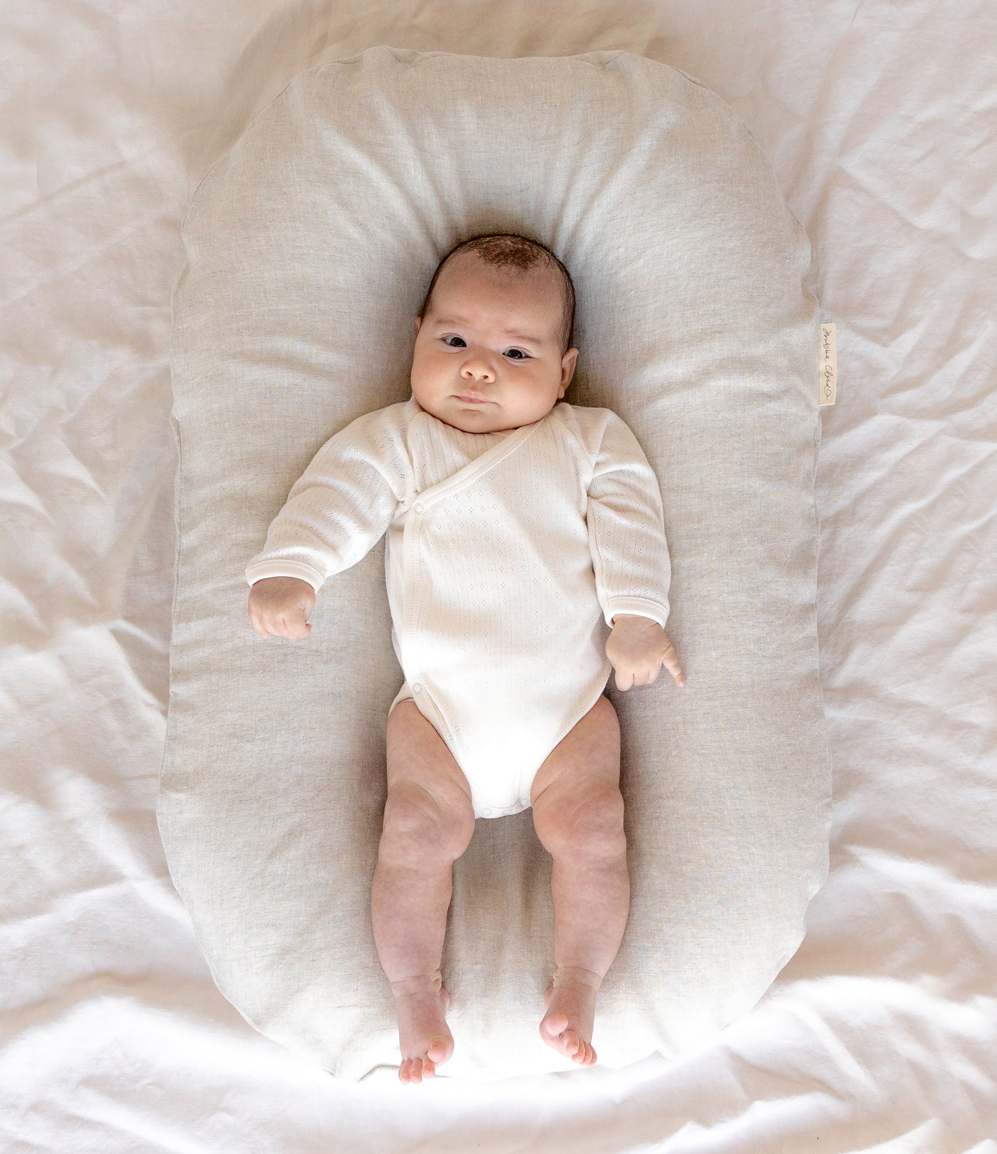 Baby Pictured In The Bubba Cloud Sand Dune Baby Lounger Wearing A White Long Sleeve White Body Suit Looking At The Camera. The Background Is A White Sheet.